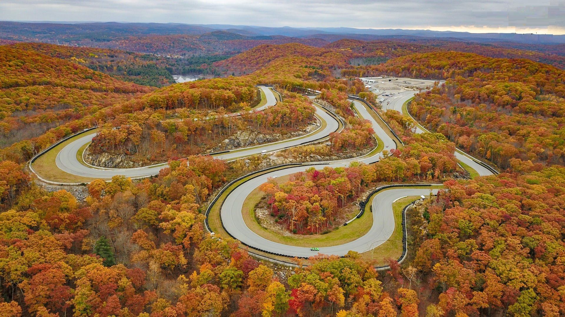 Aerial view in autumn of the track winding through hairpins and esses on a wooded hillside, with the paddock in the distance.